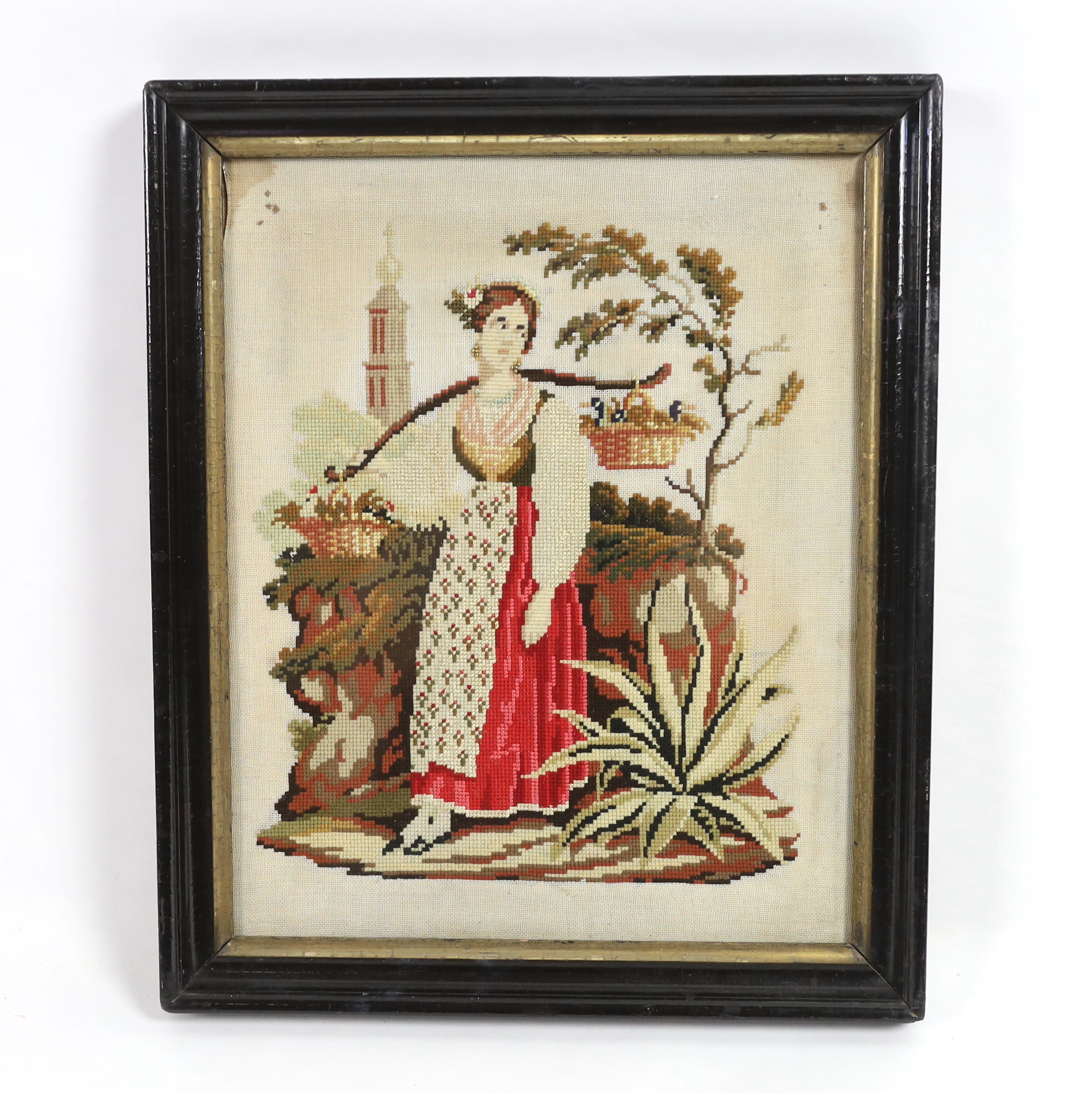 A framed 19th century Berlin wool worked embroidery of a peasant girl carrying panniers by a church, with the steeple in the background, 30cm x 38cm